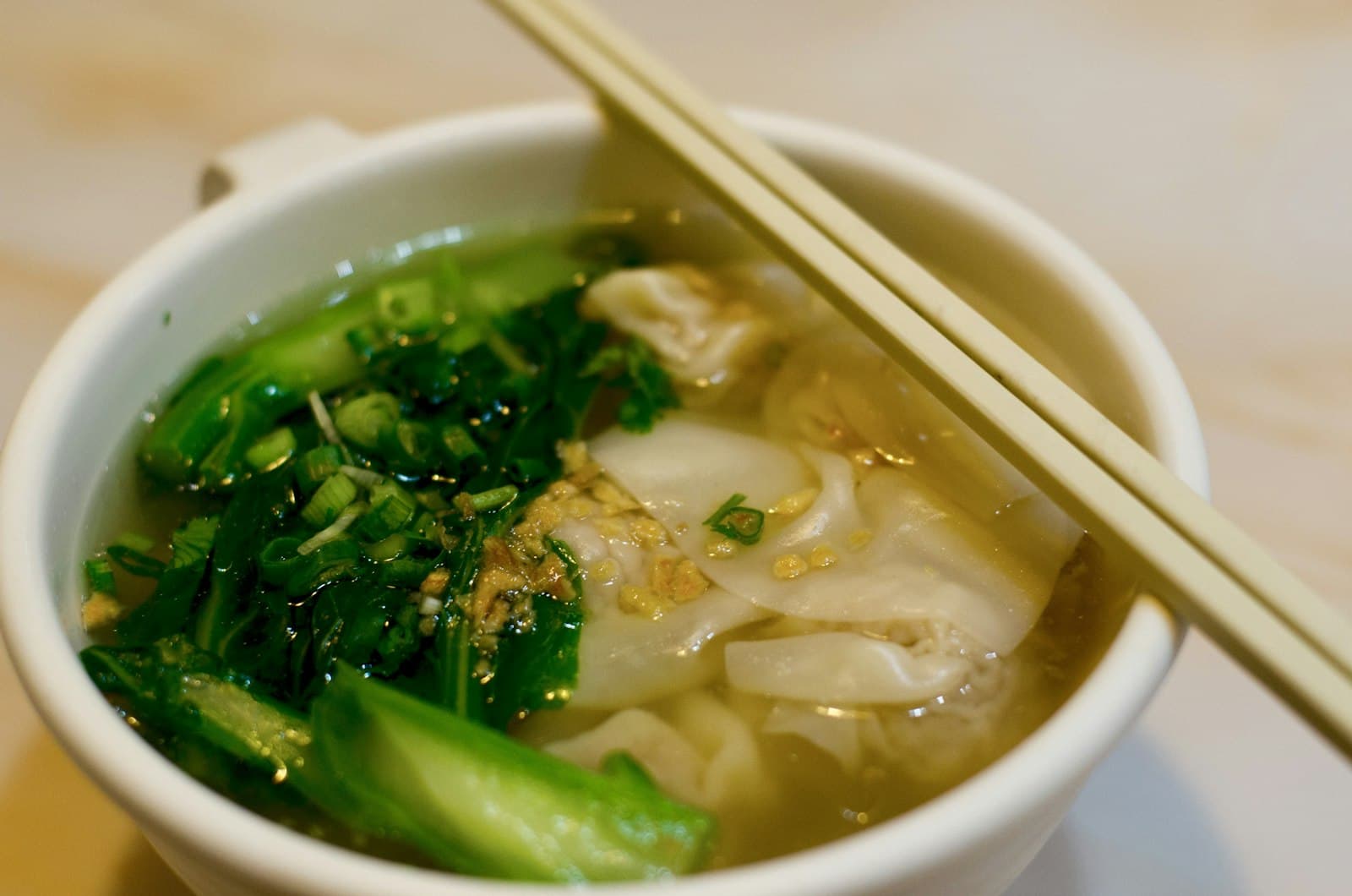 Wonton soup with leafy greens and green onions in clear broth, in a white bowl with chopsticks resting on the rim