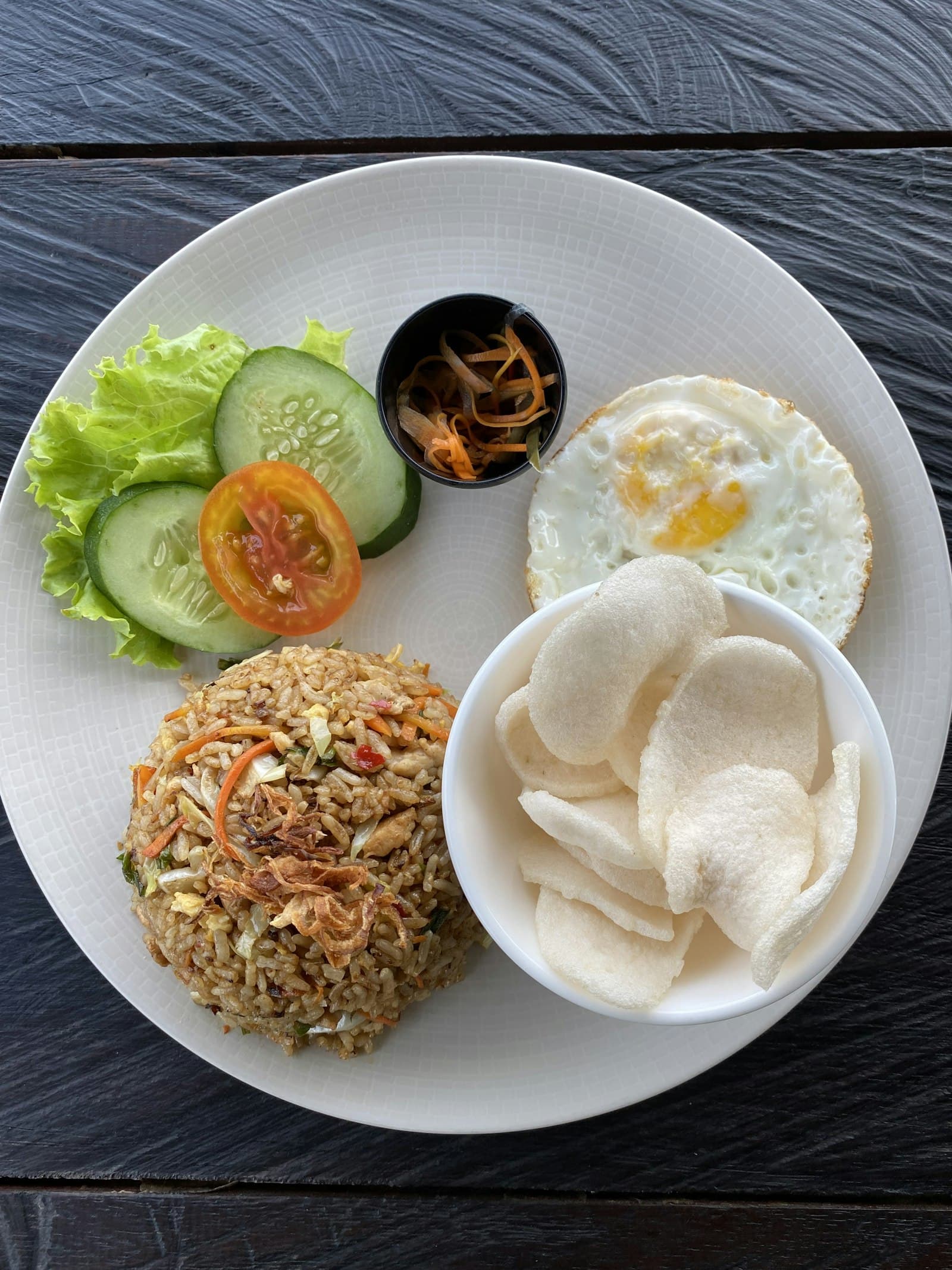 Dome of nasi goreng topped with fried shallots on a white plate with a fried egg, prawn crackers, cucumber, tomato and lettuce