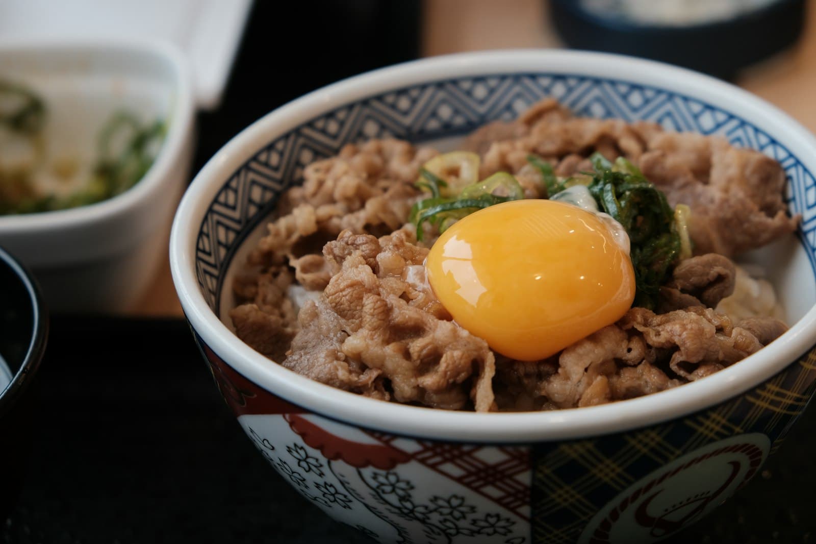 Gyudon of simmered sliced beef over rice topped with a raw egg yolk and green onions in a patterned bowl