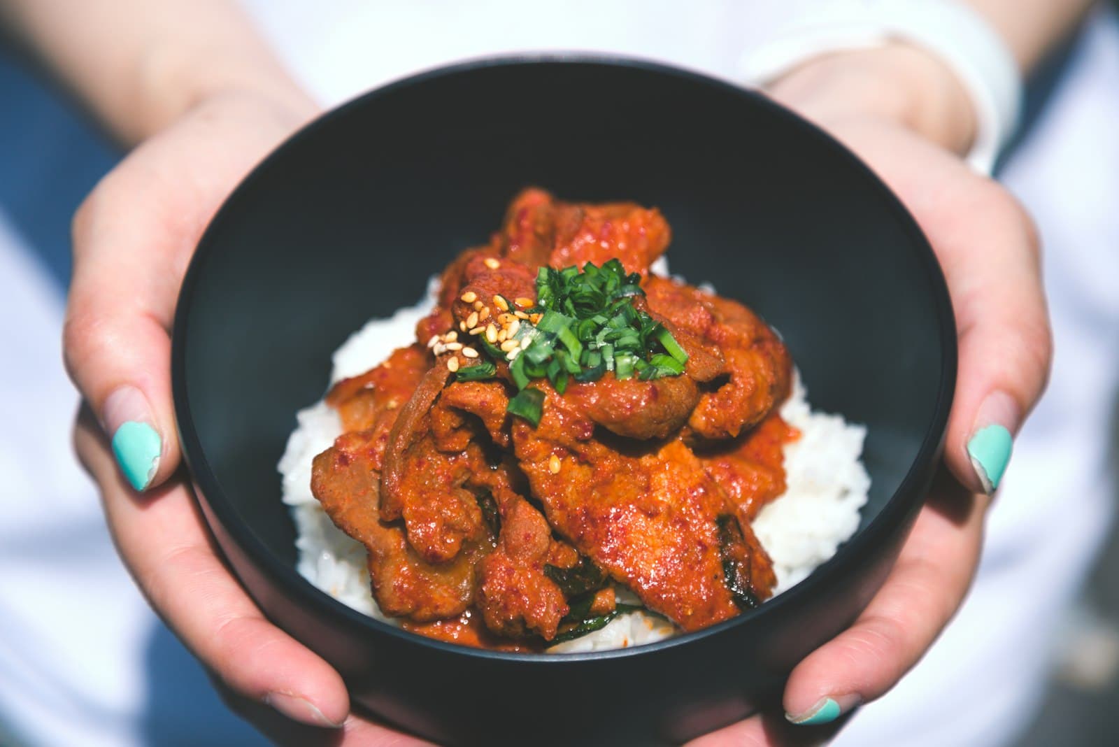 Spicy red gochujang pork over white rice in a black bowl, topped with chopped green onions and sesame seeds