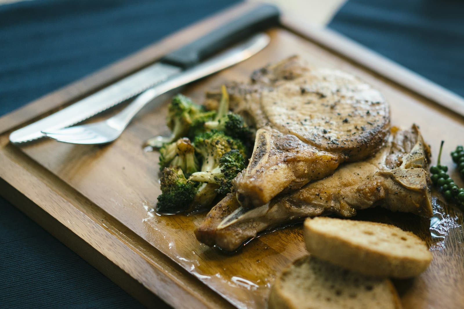 Two pepper-crusted bone-in pork chops on a wooden board with roasted broccoli and sliced bread
