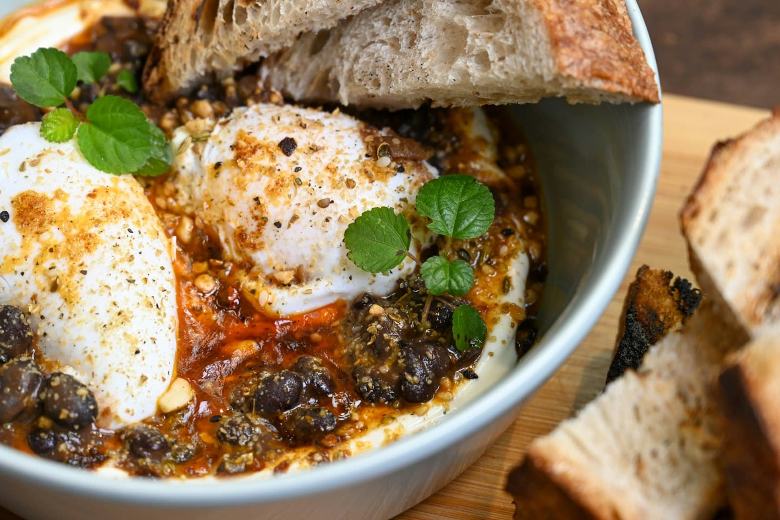 Two poached eggs on yogurt with red chili butter, crushed nuts and mint leaves, with slices of toasted sourdough