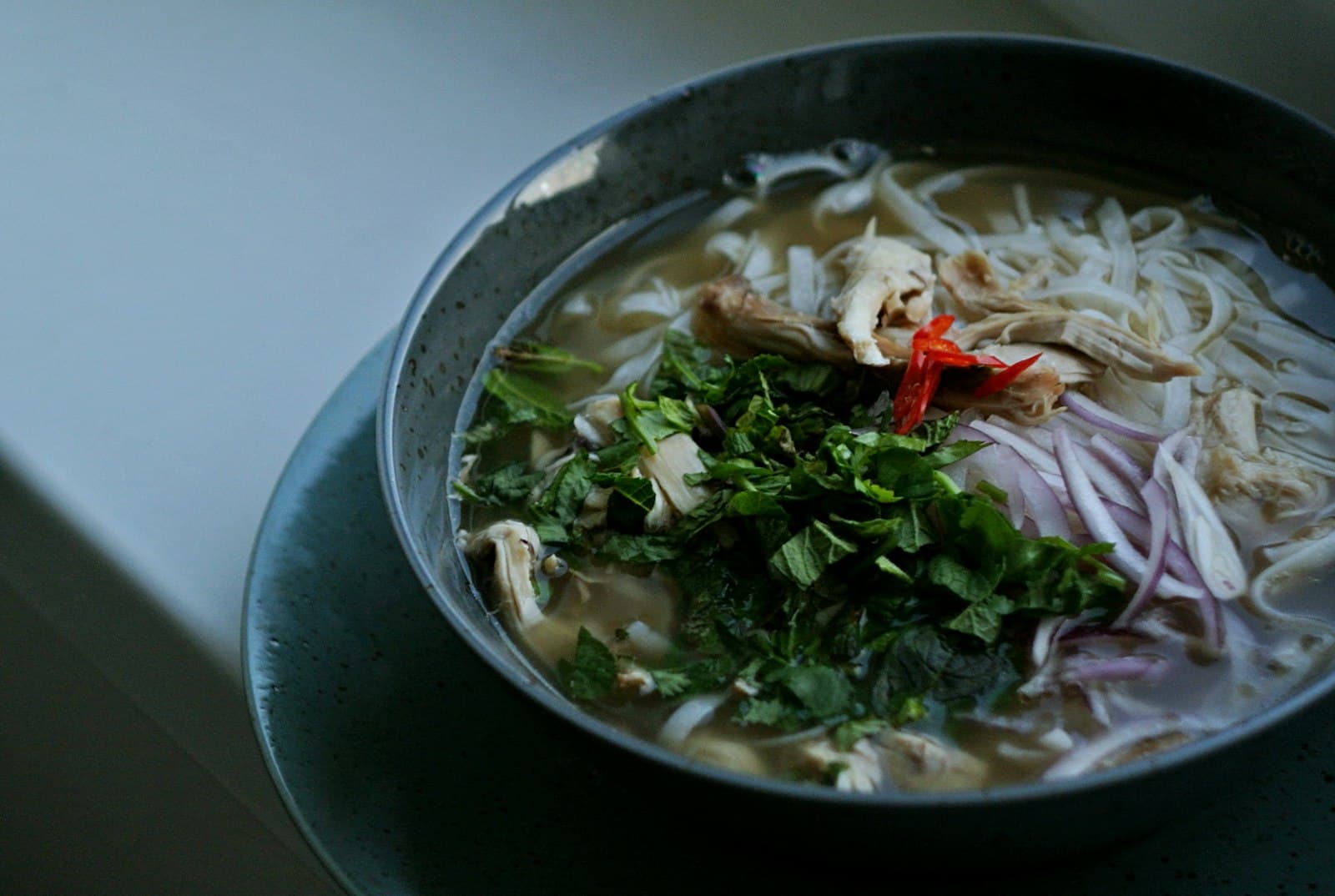 Bowl of chicken pho with rice noodles, shredded chicken, fresh herbs, sliced red onion and red chilli