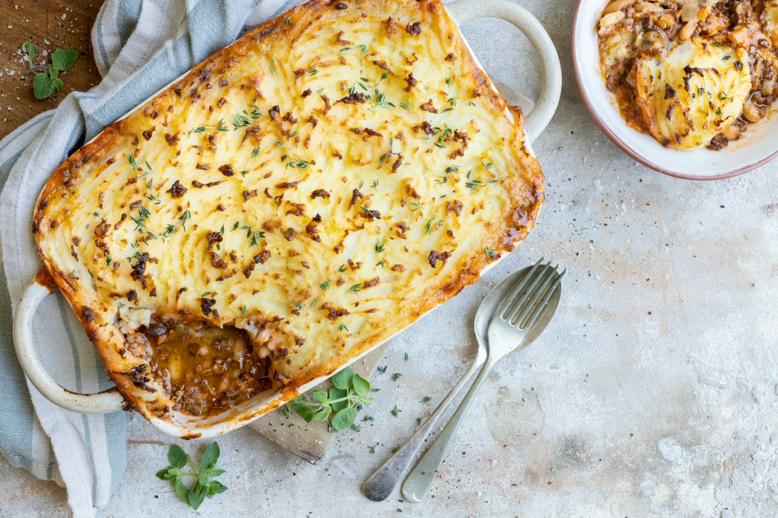 Shepherd's pie with a browned mashed potato topping and thyme in a baking dish, with one serving scooped out beside two forks
