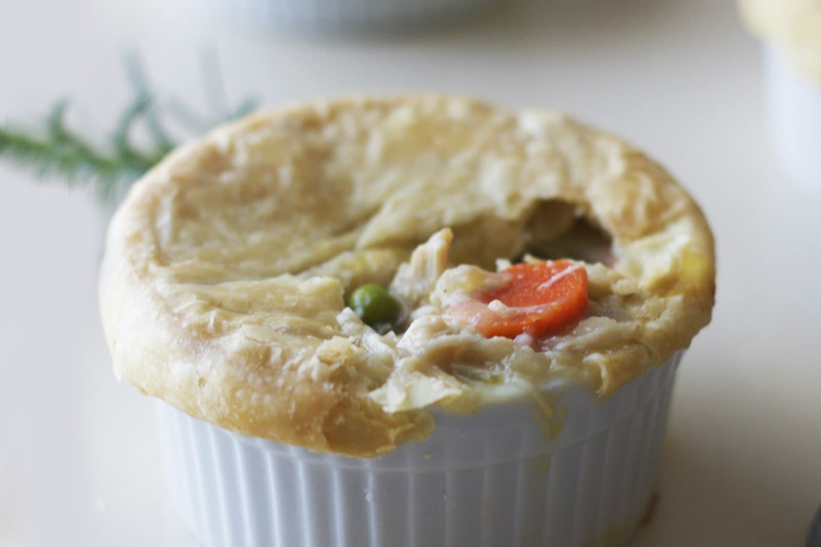 Chicken pot pie in a white ramekin with a golden crust broken open to show chicken, peas and carrots