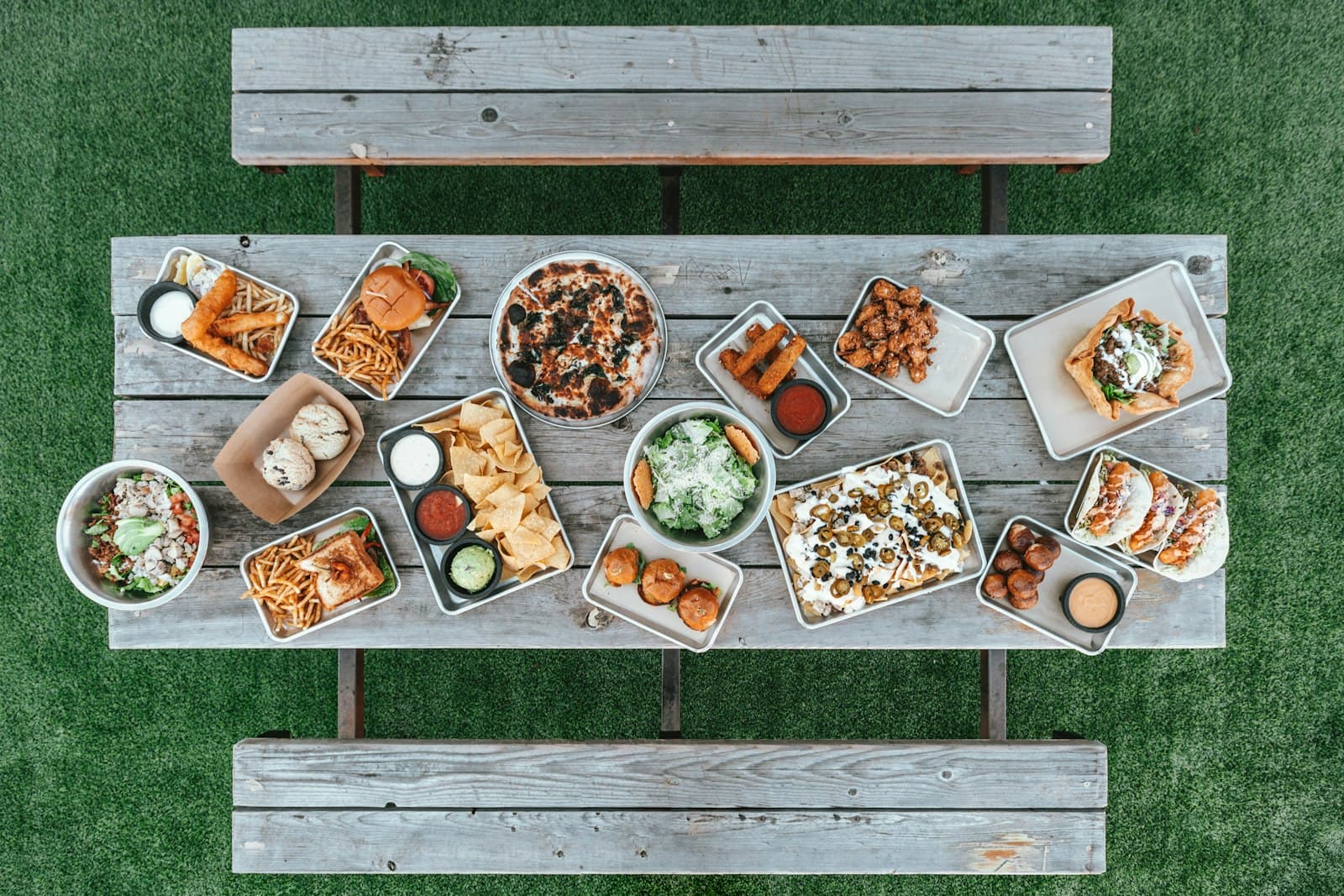 Overhead view of a wooden table covered with party food, including pizza, nachos, chips with dips, burgers and tacos