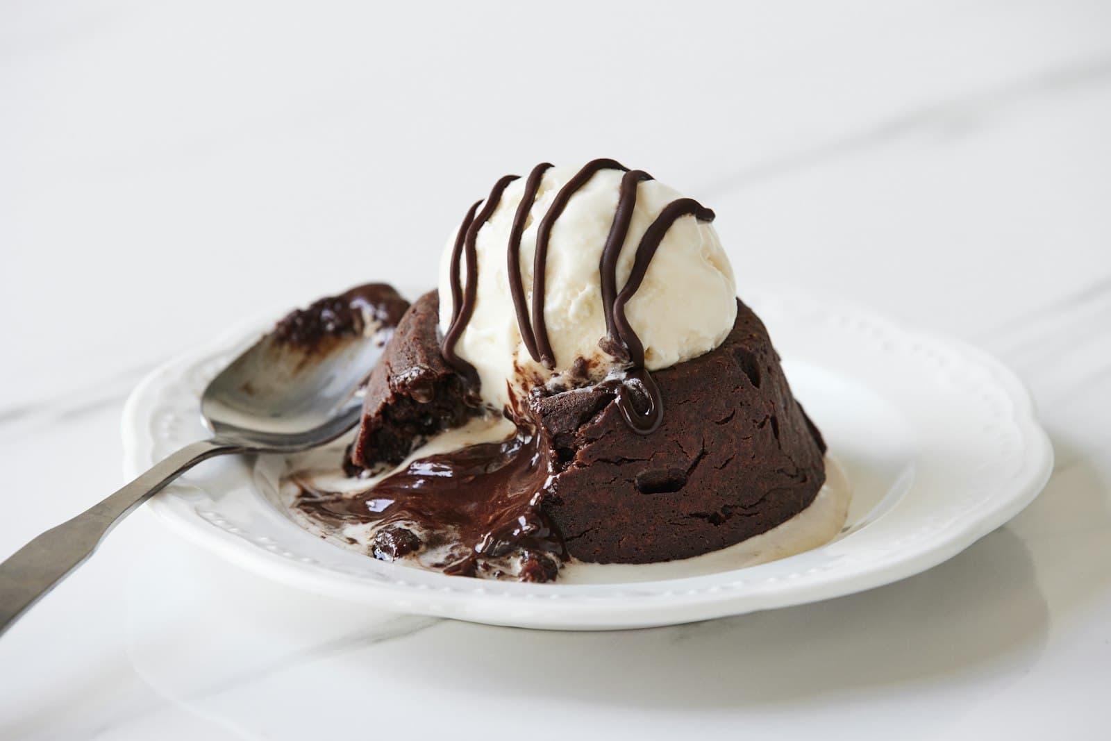 Chocolate lava cake with a molten centre, topped with vanilla ice cream and chocolate sauce, with a spoon on a white plate