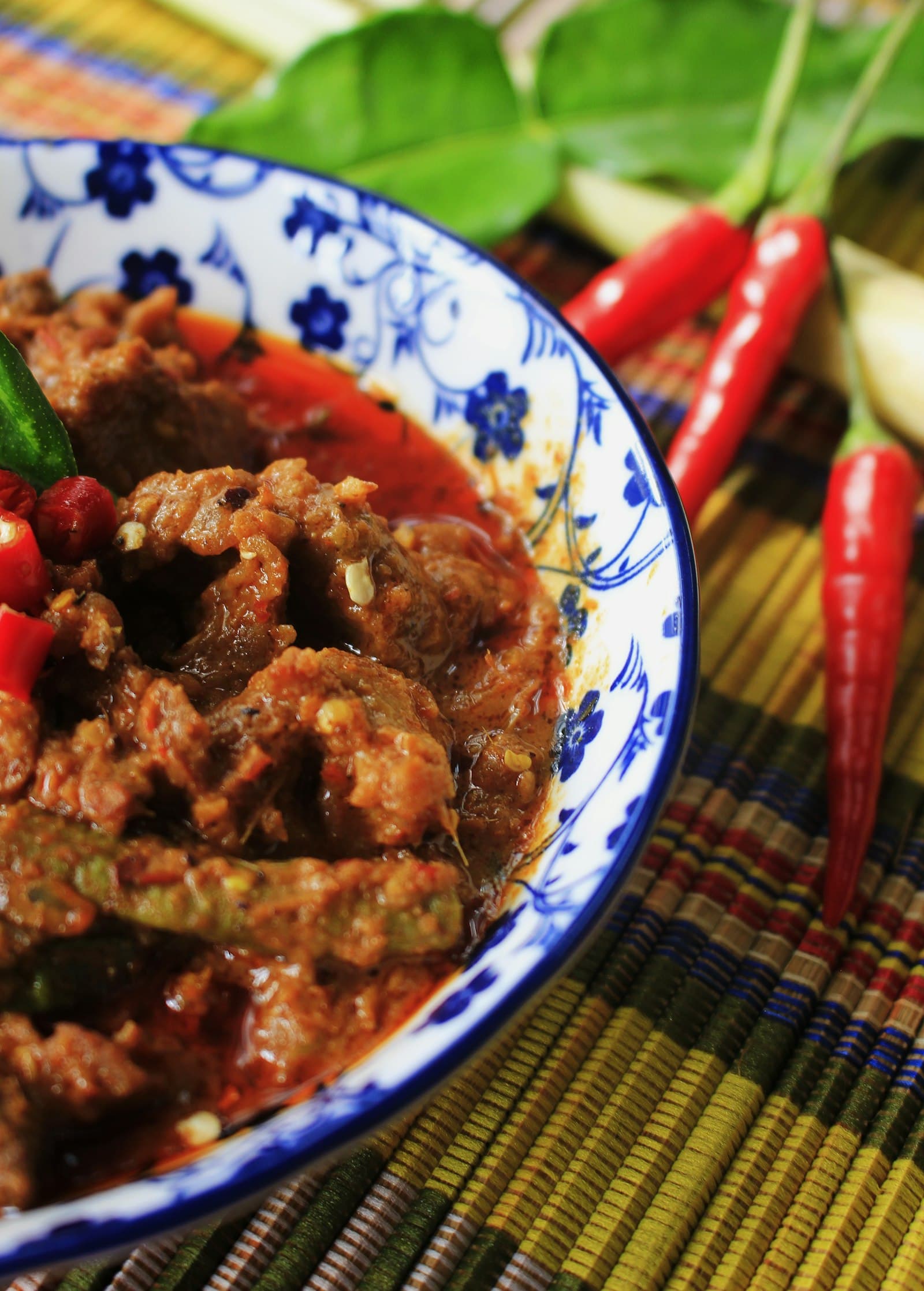 Beef rendang in a blue and white patterned bowl with fresh red chillies, lemongrass and lime leaves beside it