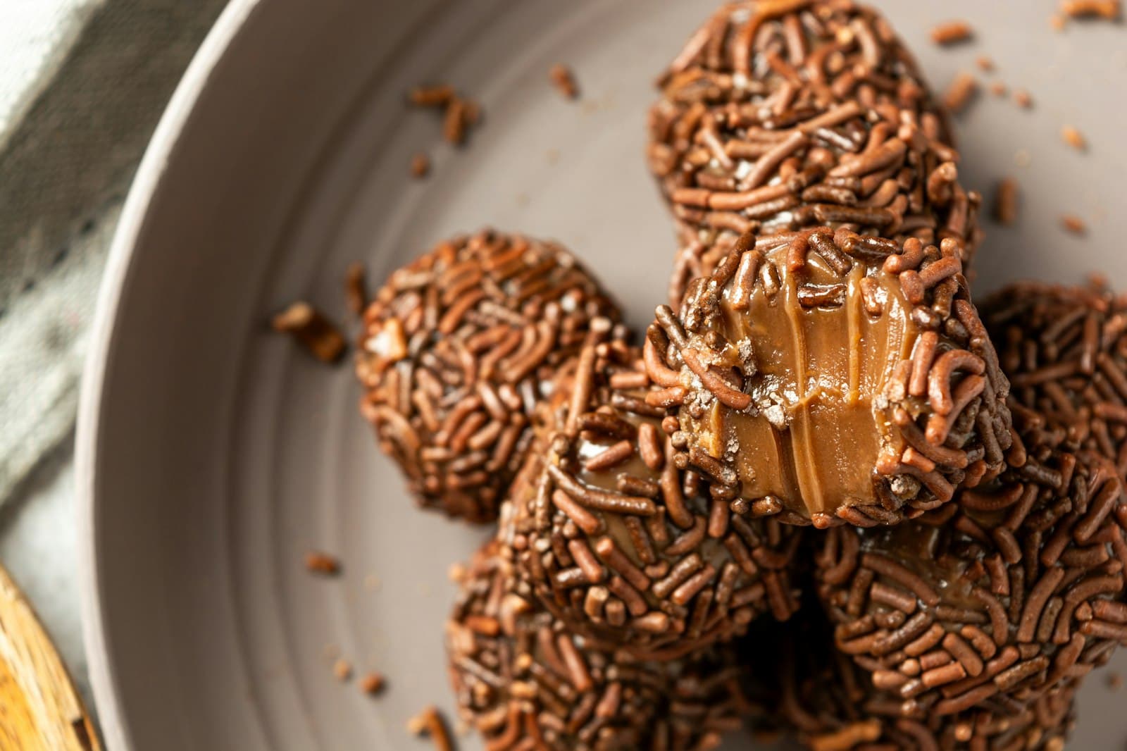 Brigadeiros coated in chocolate sprinkles on a grey plate, one bitten to show the soft fudge centre